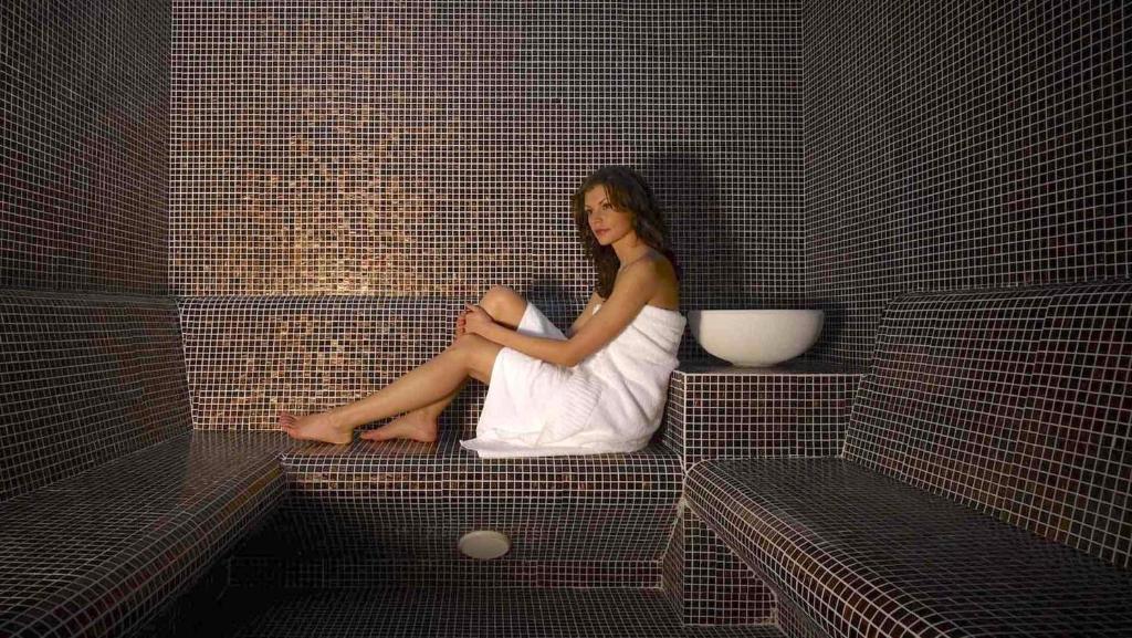 Skaista sieviete, apvīta dvielī, atpūšas mūsdienīgā tvaika pirtī ar melnām mozaīkas flīzēm. Svaiguma un relaksācijas atmosfēra.
