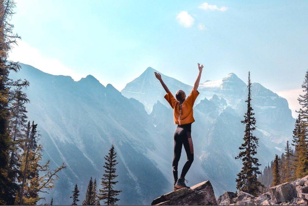Sieviete stāv kalnu virsotnē, izstiepjot rokas uz augšu, ar skaistu kalnu ainavu fonā. Gaisma ir mīksta, radot mierīgu atmosfēru.