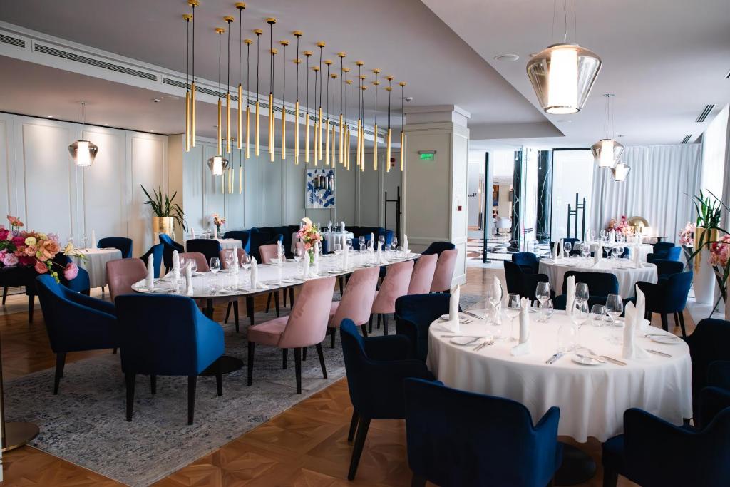 Elegants restorāns ar modernu interjeru, kurā redzami apaļi un garie galdi ar dārgiem galda piederumiem. Telpā ir gaišas krāsas un stilīgas lampas, radot patīkamu atmosfēru.