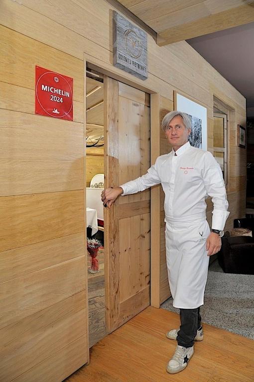 Šefpavārs stāv pie restorāna durvīm, kur redzams 2024. gada Michelin zīme. Restorāns ir eleganti iekārtots ar koka apdari.