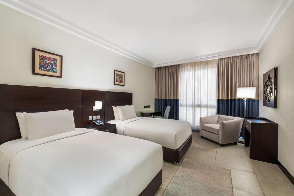 Mūsdienīga viesnīcas istaba ar divām gultām, rakstāmgaldu un ērtu atpūtas krēslu. Labi apgaismota telpa ar logiem, kas nodrošina dabīgu gaismu.