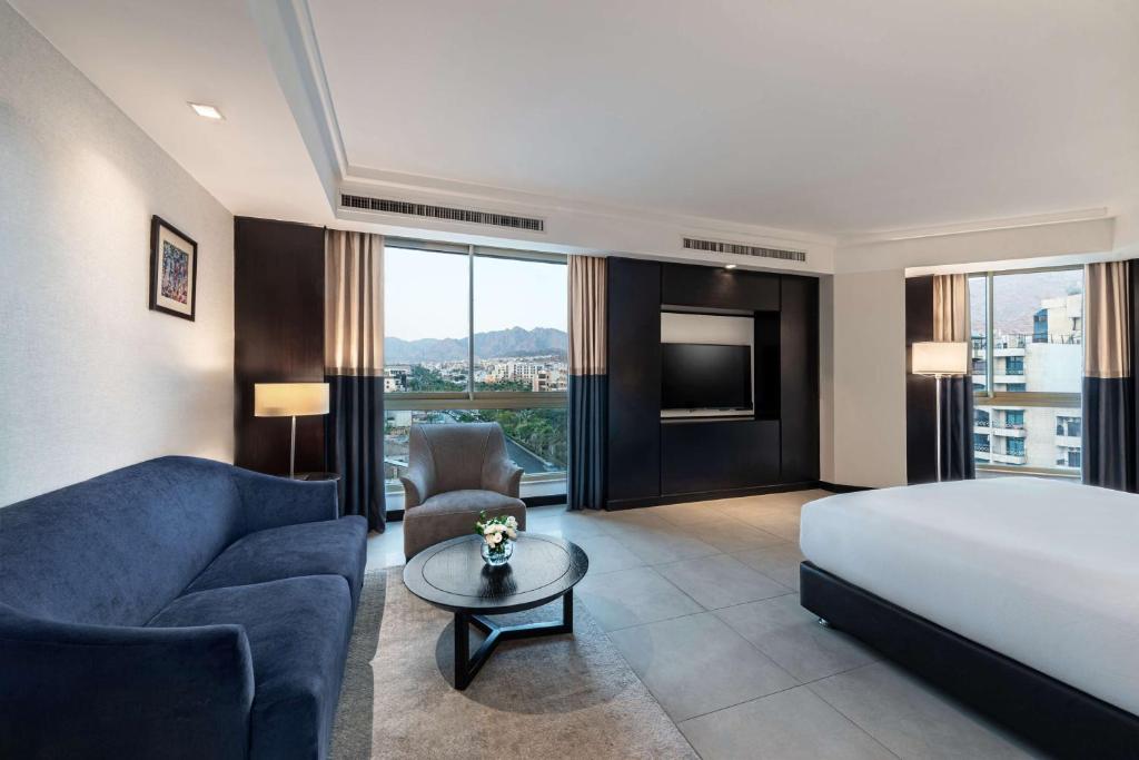 Mūsdienīga viesnīcas numura iekārtojums ar skatu uz pilsētu, iekļaujot dīvānu, krēslu un galdu. Gaismas un logi nodrošina gaišu un plašu atmosfēru.