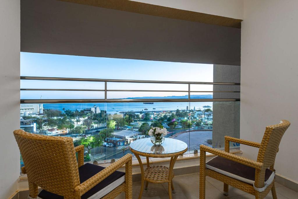 Skats no balkona uz jūru, kur divi rati sēdekļi un galds ar ziediem. Aiz loga redzams pilsētas ainava un krasta līnija.