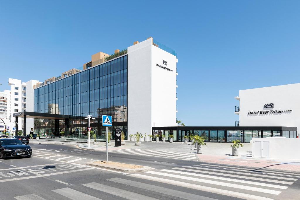 Mūsdienīga viesnīca ar lieliem stikla logiem un modernu arhitektūru, kas atrodas pie ielas. Viesnīcas nosaukums "Hotel Best Tritón" redzams uz ēkas fasādes.