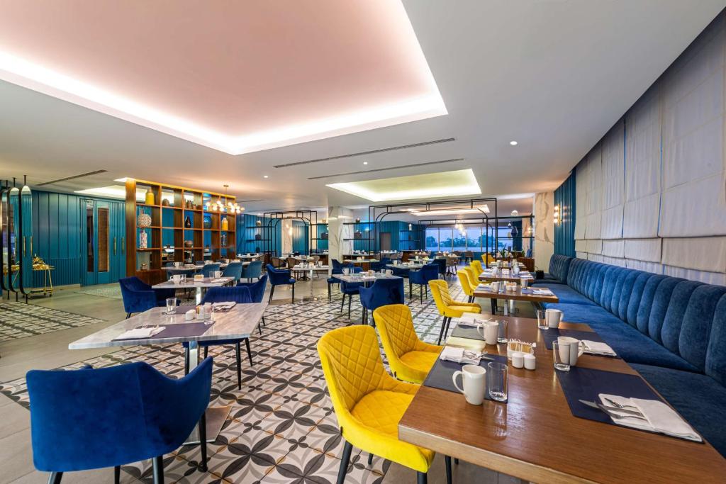 Mūsdienīgs restorāns ar stilīgu interjeru, kurā apvienoti zili un dzelteni krēsli, eleganti galdi un grezns apgaismojums, radot patīkamu atmosfēru.