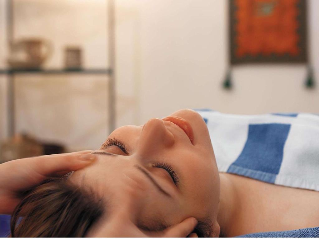 Sieviete relaksējošā masāžas sesijā ar aizvērtām acīm, izbaudot mierīgu atmosfēru. Fons ietver interjera elementus, kas rada nomierinošu noskaņu.