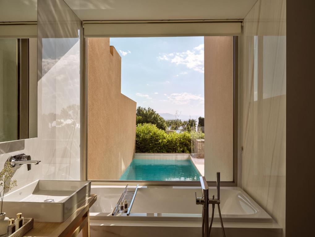 Mūsdienīgs vannas istabas interjers ar skatu uz baseinu un dārzu, nodrošinot relaksējošu atmosfēru. Dabas gaisma plūst caur lieliem logiem, radot gaišu un atvērtu telpu.