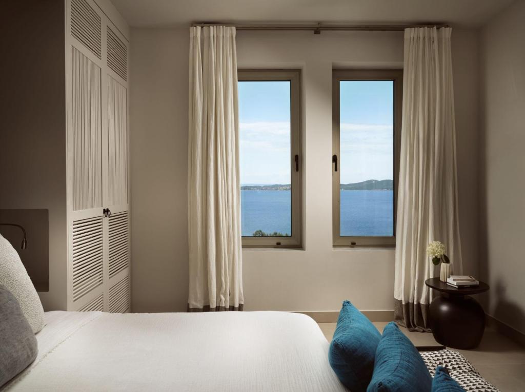 Mūsdienīgs guļamistabas interjers ar plašiem logiem, kas piedāvā skatu uz jūru. Gultā ir dekoratīvi spilveni, bet blakus ir neliels galdiņš ar ziedu kompozīciju.