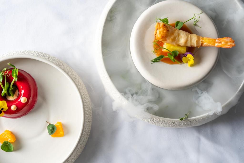 Elegants un māksliniecisks ēdiens uz baltas galdautas. Uz vienas plāksnes ir sarkans deserts ar ziediem un augļiem, bet uz otras – garnelē ietinēta uzkoda ar krāsainiem dārzeņiem.