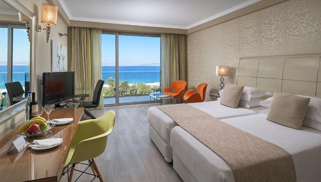 Mūsdienīga viesnīcas numura iekārtojums ar skatu uz jūru, divām gultām, ērtu dīvānu un galdu ar augļiem. Lieliskas gaismas un gaišas krāsas rada patīkamu atmosfēru.