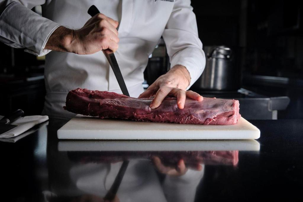 Šefpavārs, izmantojot nazi, sagriež gaļu uz griešanas dēļa, izceļot precizitāti un profesionālu pieeju.