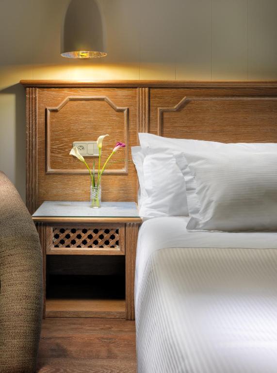 Mūsdienīgs guļamistabas interjers ar koka gultas galvu, baltiem spilveniem un ziedu pušķi uz naktsgaldiņa. Siltā apgaismojuma atmosfēra rada mierīgu un relaksējošu vidi.