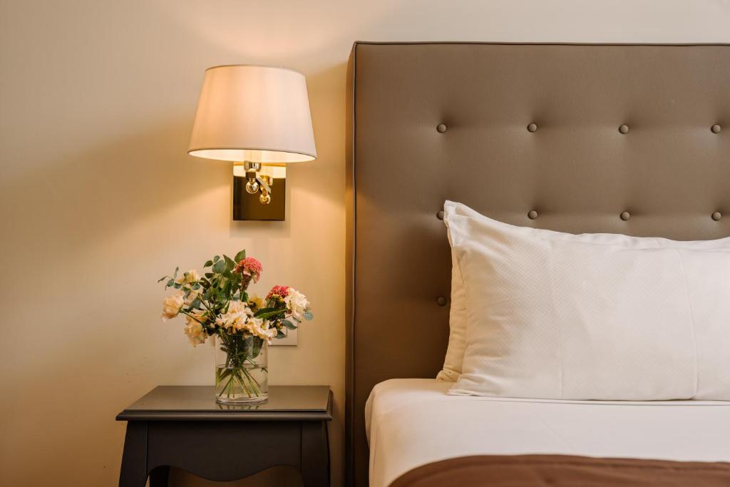 Mūsdienīgs guļamistabas interjers ar ērtu gultu un dekoratīvu lampu. Uz naktsgaldiņa atrodas ziedu pušķis, kas piešķir siltumu un eleganci telpai.