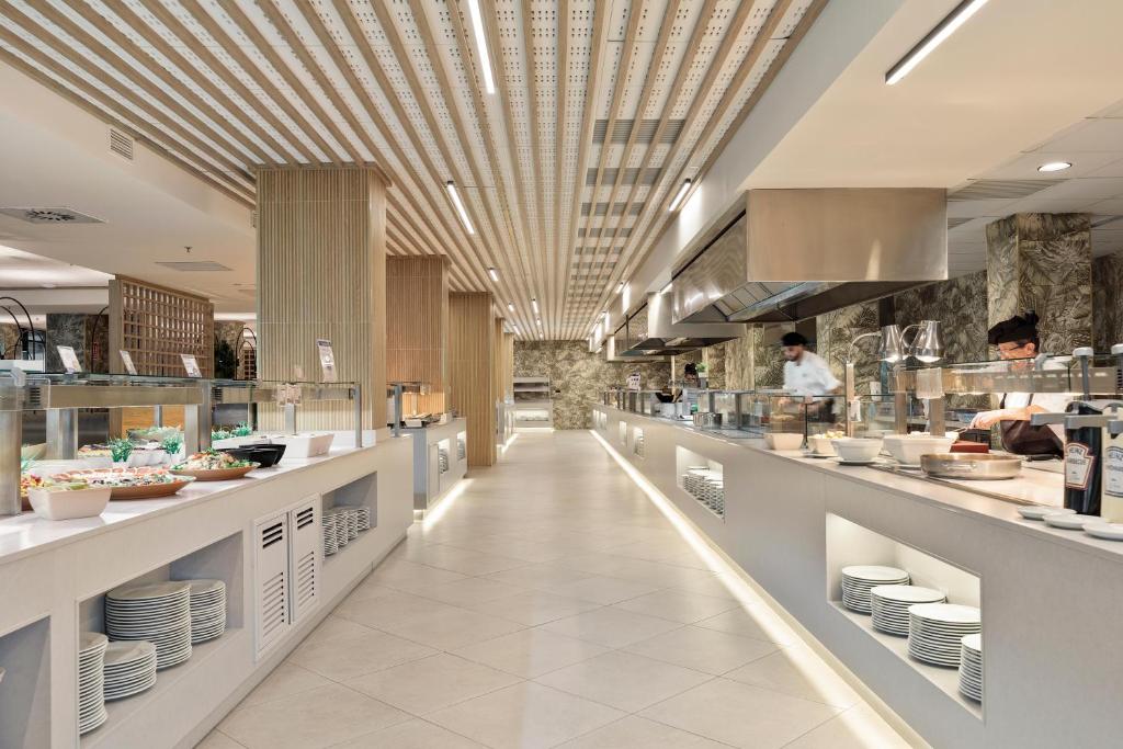 Mūsdienīgs bufetes restorāns ar elegantu interjeru, kurā redzami ēdieni un šefpavāri, kas strādā aiz bāra. Gaismas un koka elementi rada patīkamu atmosfēru.