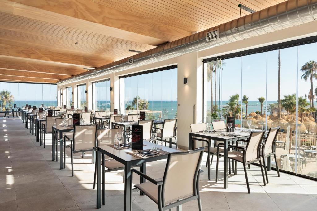 Mūsdienīgs restorāns ar plašām logu sieniņām, kas piedāvā skatu uz jūru un palmām, radot relaksējošu atmosfēru. Galdi ir eleganti sakārtoti ar ēdienkartēm un glāzēm.