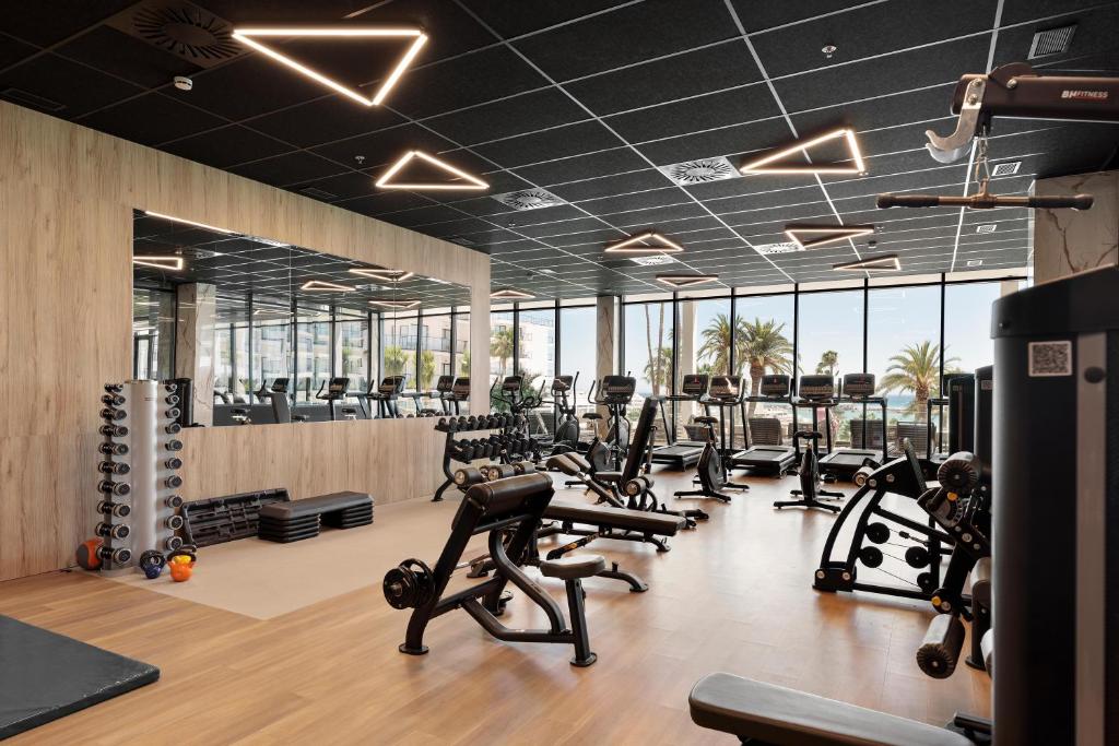 Mūsdienīgs sporta zāles iekārtojums ar dažādām trenažieru iekārtām un svaru komplektiem, kas nodrošina lielisku treniņu pieredzi. Lielie logi piedāvā skatu uz ārpusi ar palmām un sauli.