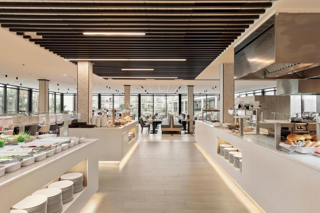 Mūsdienīgs restorāna interjers ar plašām logiem, gaišām krāsām un dažādām ēdienu iespējām uz bufetes galda. Apmeklētāji var baudīt ērtu un stilīgu ēdināšanas pieredzi.