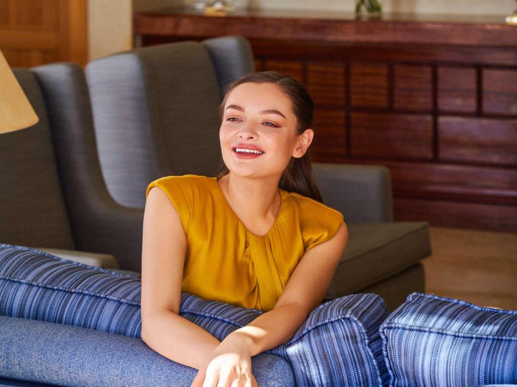 Sieviete ar priecīgu smaidu sēž uz dīvāna, tērpta dzeltenā blūzē, viesmīlīgā telpā ar mīkstiem dīvāniem.