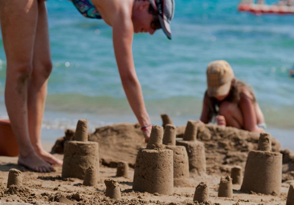 Divas meitenes spēlē smilšu pilis pludmalē, viena no viņām pievērš uzmanību savam smilšu veidojumam, kamēr otra sēž tuvumā. Zils ūdens un saulaina laika atmosfēra rada priecīgu noskaņu.