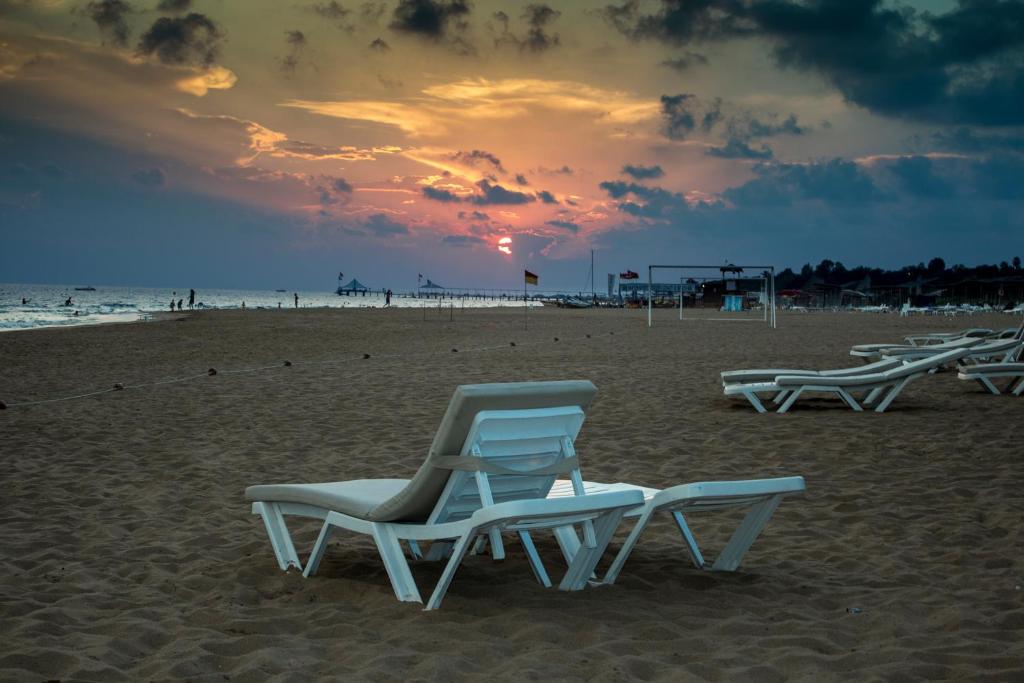 Pludmales ainava ar baltām sauļošanās krēslām un saulrietu, kas rada mierīgu atmosfēru. Aiz krēsliem redzama jūras un debesu krāsu spēle.