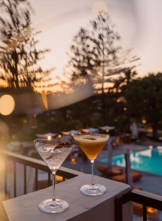 Divi kokteiļi, viens martini un viens dzeltens kokteilis, novietoti uz balkona ar skatu uz peldbaseinu un saulrietu. Apkārtējo ainavu papildina koki un mīksta apgaismojums.