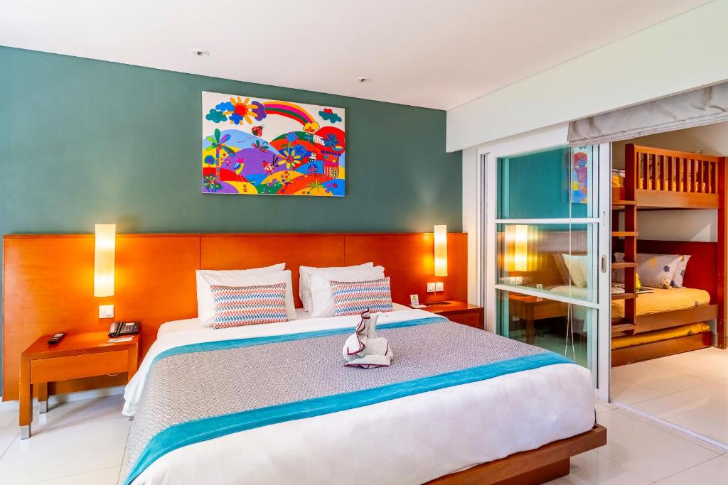 Mūsdienīgs viesnīcas numurs ar divguļamo gultu, bērnu gultu un krāsainu mākslas darbu uz sienas. Interjers ir gaišs un draudzīgs, ar mūsdienīgām mēbelēm un komfortablu atmosfēru.