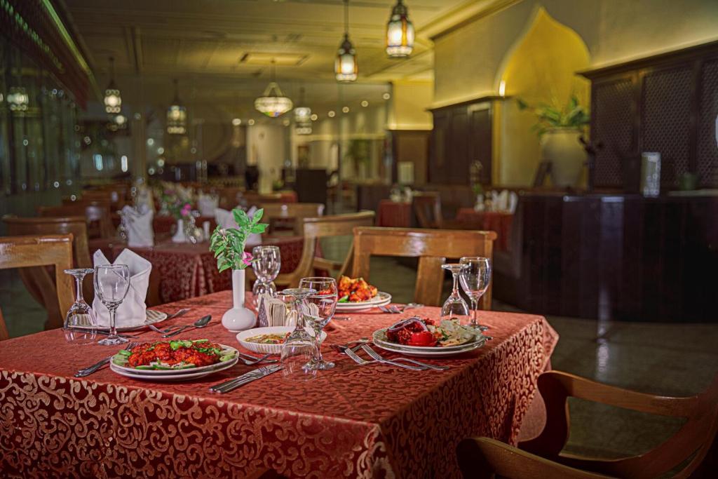 Elegants restorāns ar sarkanu galdautu, izrotātu ar ēdieniem un glāzēm, radot viesmīlīgu atmosfēru. Detaļas iekļauj ziedus un apgaismojumu, kas piešķir siltu noskaņu.