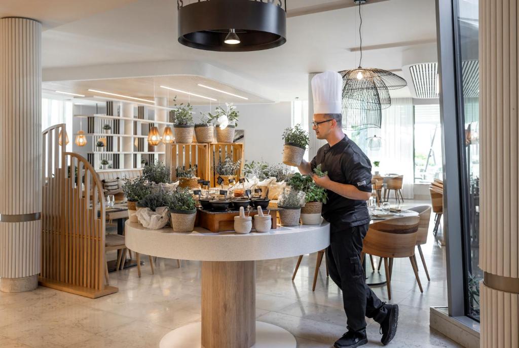 Šefpavārs, tērpies melnā uniformā un cepurē, strādā modernā restorānā, kārtojot augus uz apaļā galda. Apkārt redzami eleganti ēdamgaldi un mūsdienīga interjera elementi.