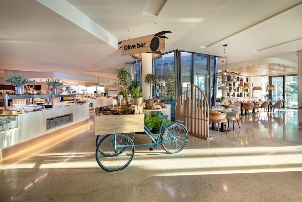 Mūsdienīga restorāna interjers ar gaisīgu dizainu, kurā redzams olīvu bārs un dekoratīvs velosipēds ar augiem. Gaismas un telpas harmonija rada patīkamu atmosfēru.