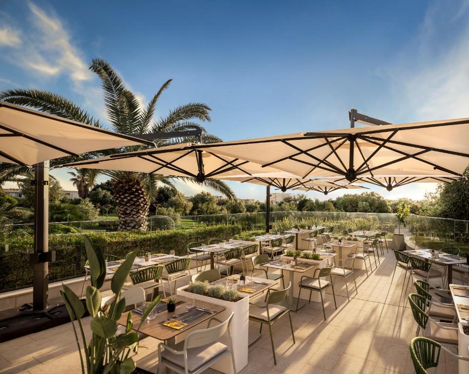 Āra restorāna terase ar elegantiem galdiem un krēsliem, apņemta ar palmām un zaļumiem, nodrošina patīkamu vidi maltītēm saulainā dienā.