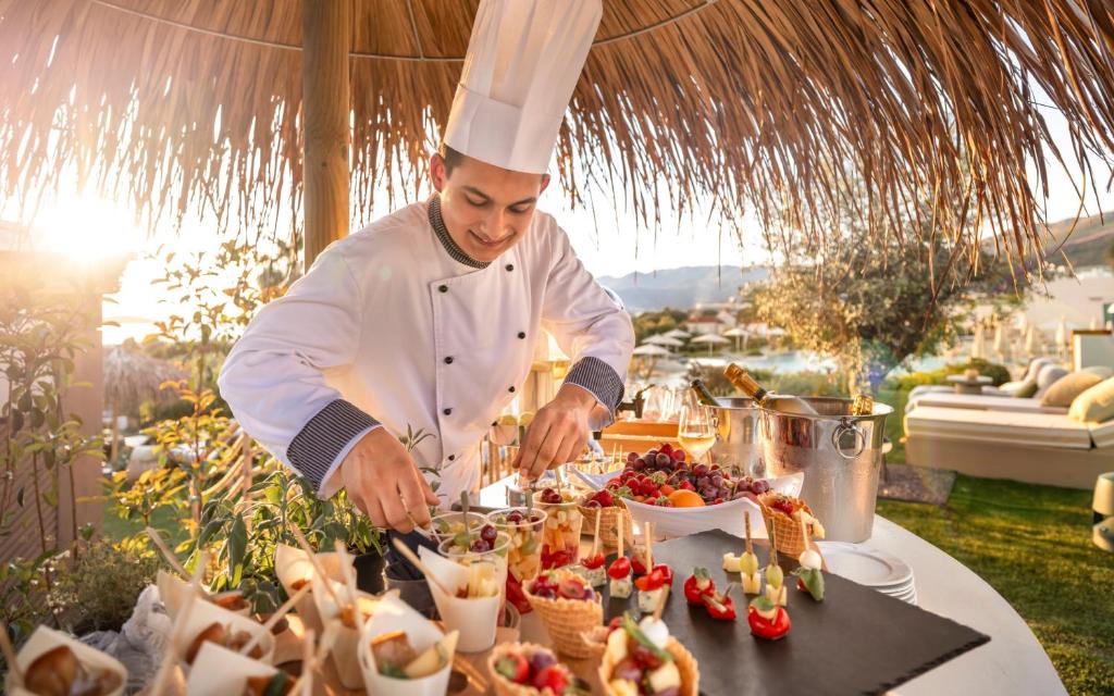 Šefpavārs gatavo gardumus saulrieta gaismā, izmantojot svaigus augļus un dažādus ēdienus uz elegantas galda. Fons ir izsmalcināta āra vide ar zāli un atpūtas zonām.