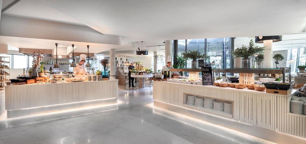 Moderns restorāns ar bufeti, kurā redzami šefpavāri, gatavojot ēdienus un piedāvājot svaigus produktus. Gaisīgs interjers ar dabīgiem elementiem un elegantu dizainu.