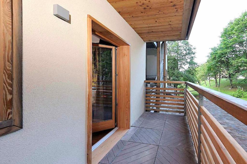 Mūsdienīgs balkons ar koka margām un durvīm, kas ved uz iekšpusi. Apkārtne ir zaļa un dabiska, radot relaksējošu atmosfētu.