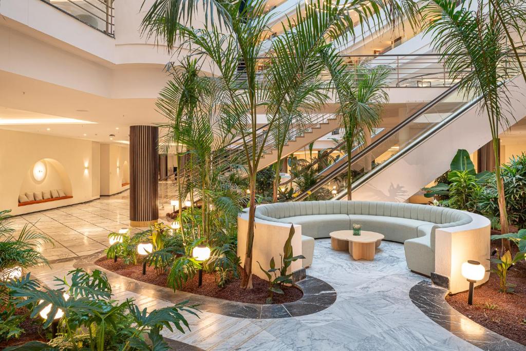Mūsdienīgs iekštelpu stūrītis ar zaļām palmām un elegantu sēžamo zonu, kurā ir apaļs galds un apgaismojums. Interjers apvieno dabīgas elementus un modernu dizainu.