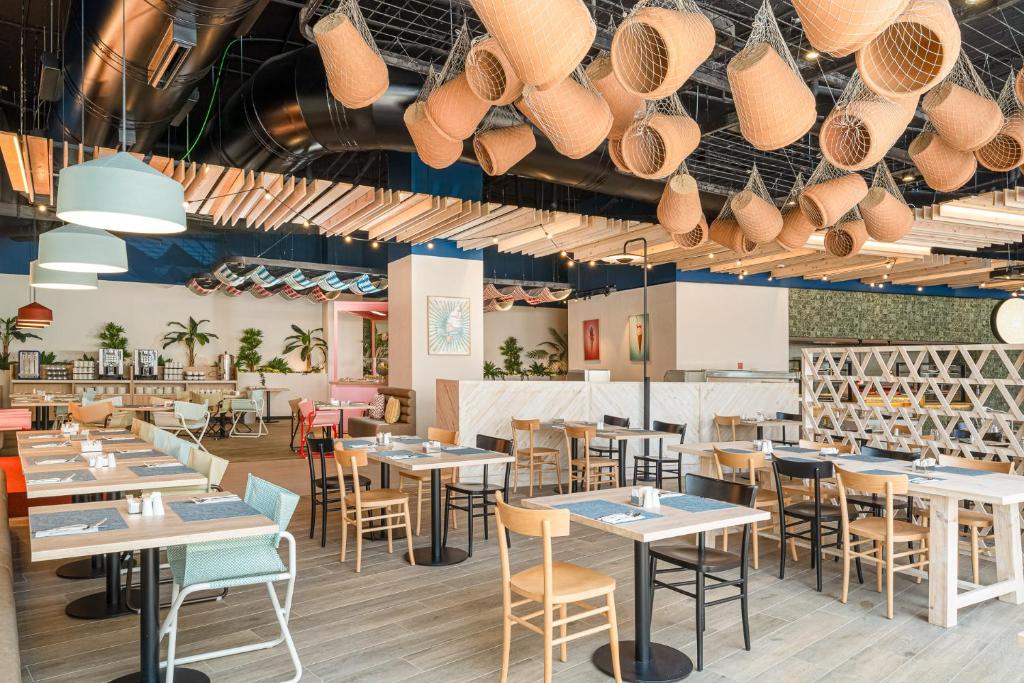 Moderns restorāns ar gaišu interjeru, kurā ir koka mēbeles un stilīgas piekārtnes. Telpā ir plaši galdi un augi, radot patīkamu atmosfēru.
