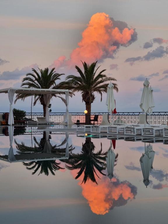 Pūķainā mākoņu atspulgs uz mierīga baseina, ap kuru aug palmas un ir redzamas saules gaismas atspīdumi. Rietošās saules krāsas radītas burvīga atmosfēra.