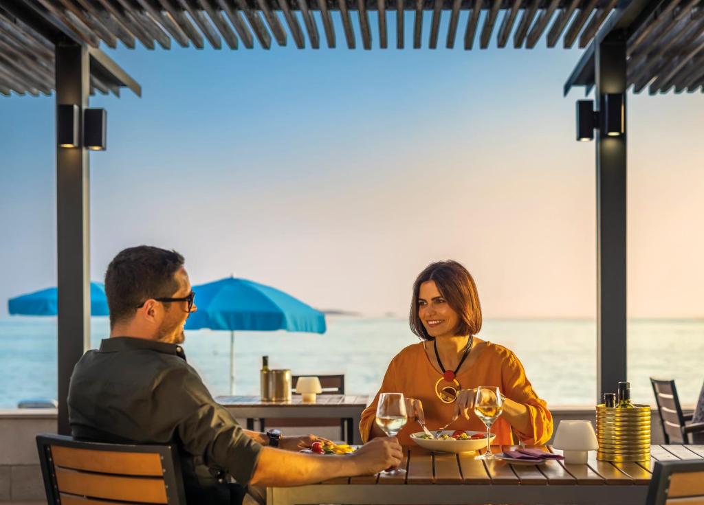 Romantiska vakariņas pie jūras, kur pāris bauda maltīti, kamēr saulriets izgaismo ainavu. Galda klājums ar vīna glāzēm un gardiem ēdieniem rada patīkamu atmosfēru.
