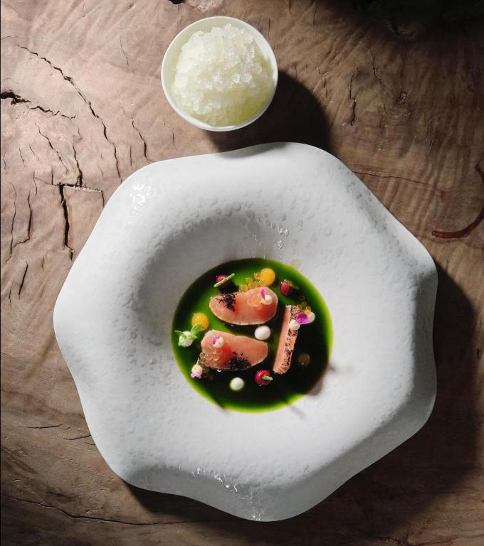 Mūsdienīga ēdiena kompozīcija ar zaļo zupu un dekoratīvām sastāvdaļām, pasniegta unikālā traukā. Blakus atrodas saldēta desertu porcija.