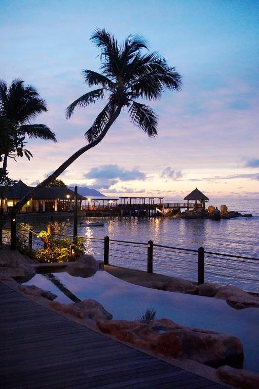 Klusā pludmales aina ar palmu un saulrietu, kas atspoguļojas ūdenī, radot mierīgu atmosfēru. Dēļu celiņš ved uz koka piestātni, kur redzamas ēkas un ainaviskas akmeņu formas.