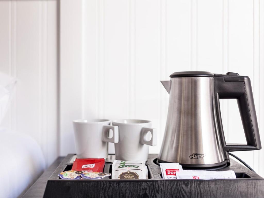 Tējkanna un divas baltas krūzes uz galda, kopā ar tējas un kafijas maisījumiem, radot mājīgu un relaksējošu atmosfēru.