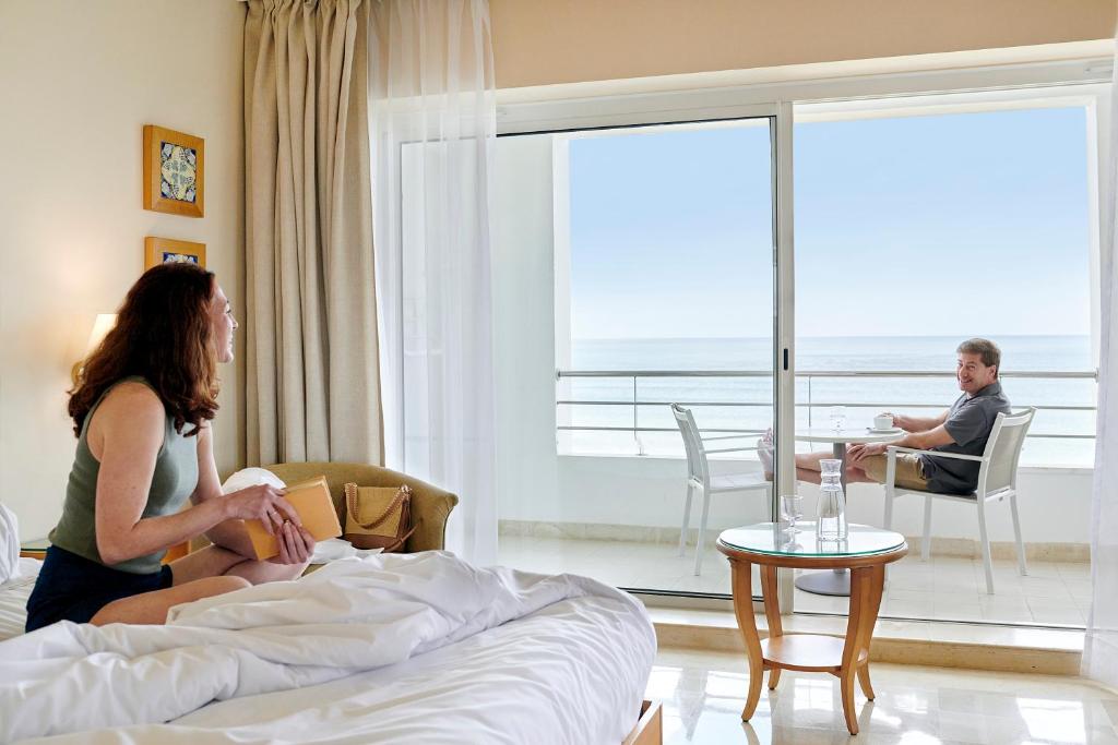 Gaismas piepildīta viesnīcas numura aina ar pāri, kas bauda rītu. Sieviete sēž gultā ar grāmatu, bet vīrietis, sēžot uz balkona, dzer kafiju un skatās uz jūru.