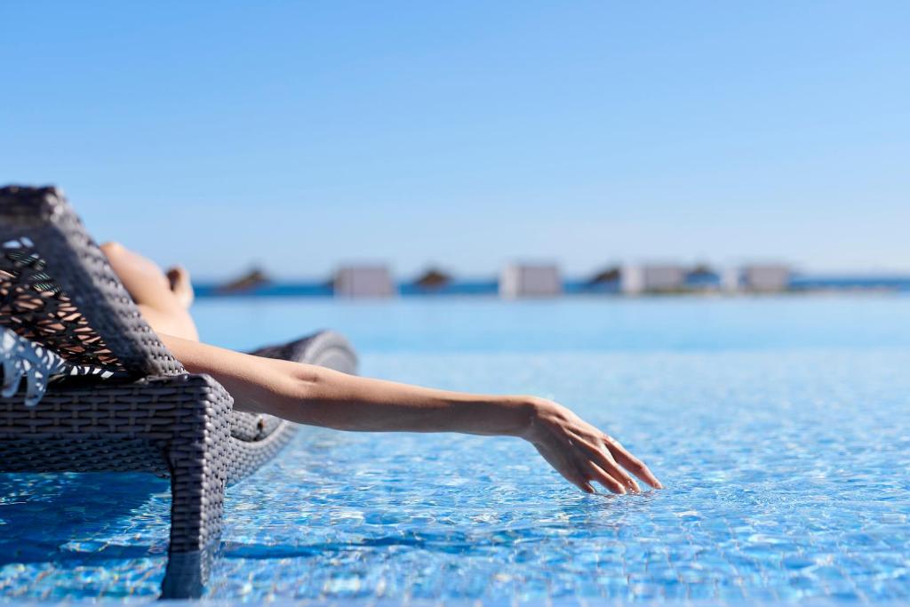 Atpūta pie baseina, roka nedaudz iemērkta ūdenī, skats uz skaidru zilo debesu un ūdens virsmu.