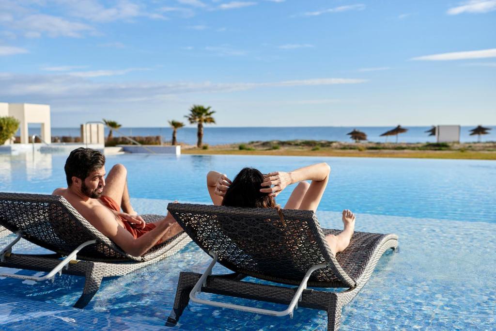 Pāris atpūšas pie baseina, baudot sauli un jūras skatu. Apkārt ir palmas un mierīga atmosfēra.