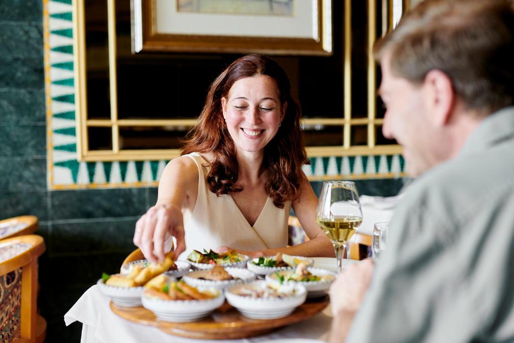 Sieviete ar brūniem matiem smaidot izvēlas ēdienu no šķīvjiem, kamēr vīrietis sēž pretī. Galda centrā ir dažādi ēdieni un glāze balto vīnu.