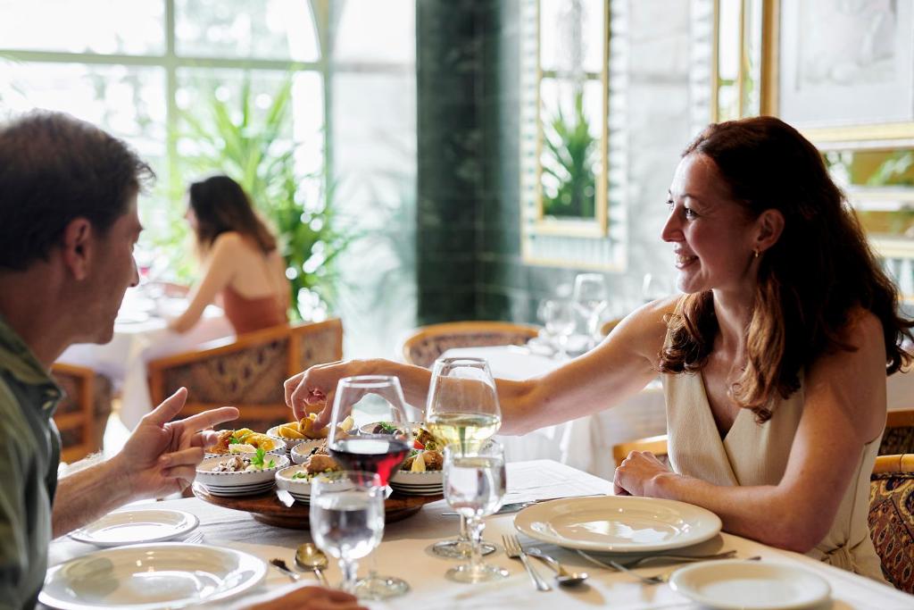 Romantisks vakariņu brīdis restorānā, kur pāris dalās gardēžu ēdienos un bauda dzērienus. Galda centrā ir krāšņs ēdienu pasniegšanas trauks.