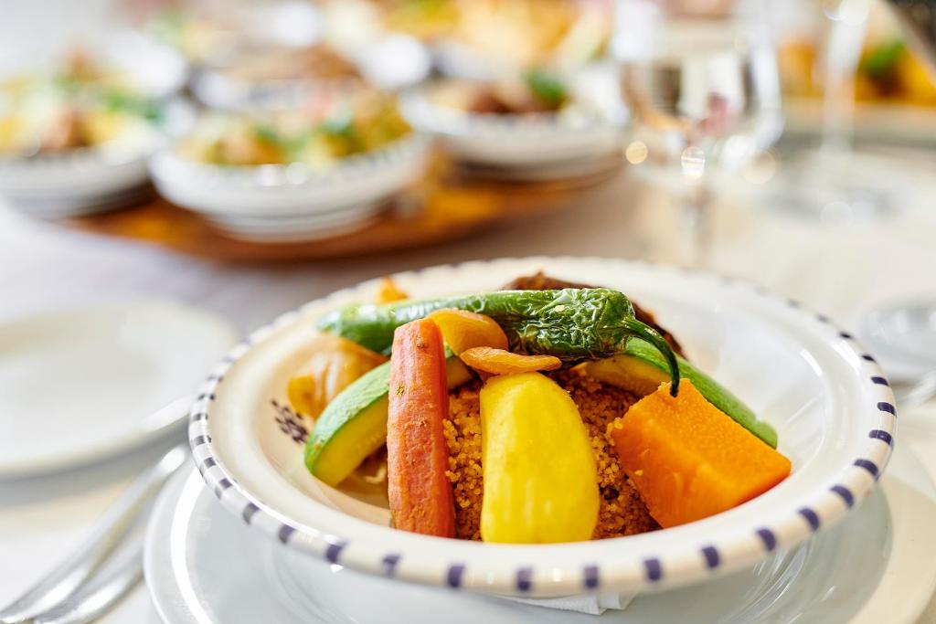 Veselīgs un krāsains ēdiens ar kuskusu, dārzeņiem un riekstiem, pasniegts uz baltas šķīvja. Fons ir izsmalcināta galda klāšana ar citiem ēdieniem.