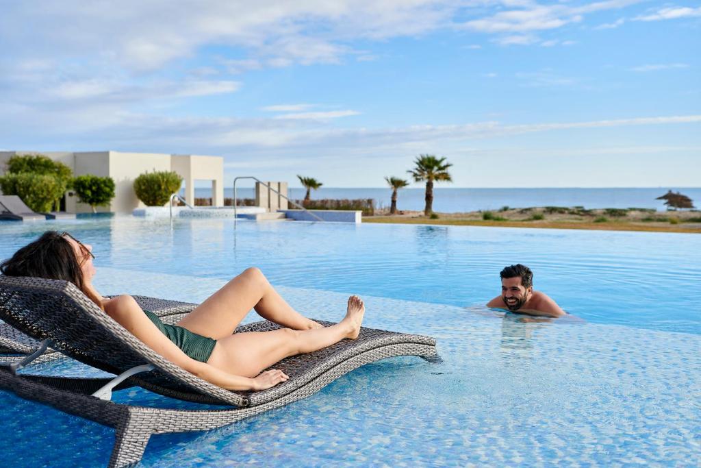 Pāris atpūšas pie baseina, kamēr viens cilvēks guļ uz šūpuļkrēsla, bet otrs peld ūdenī. Skats uz okeānu un palmu kokiem veido relaksējošu atmosfēru.