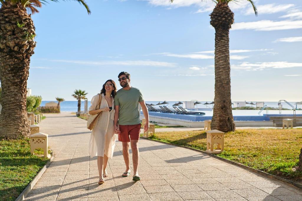 Pāris pastaigā gar jūru, saulainā dienā, starp palmām un zālāju. Romantiska atmosfēra, kas iedvesmo brīvdienu noskaņu.