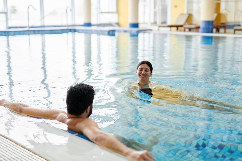 Divi cilvēki peld baseinā, viens atpūšas uz malas, bet otrs peld. Ūdens ir skaidrs un gaišs, telpa ir gaiša un moderna.