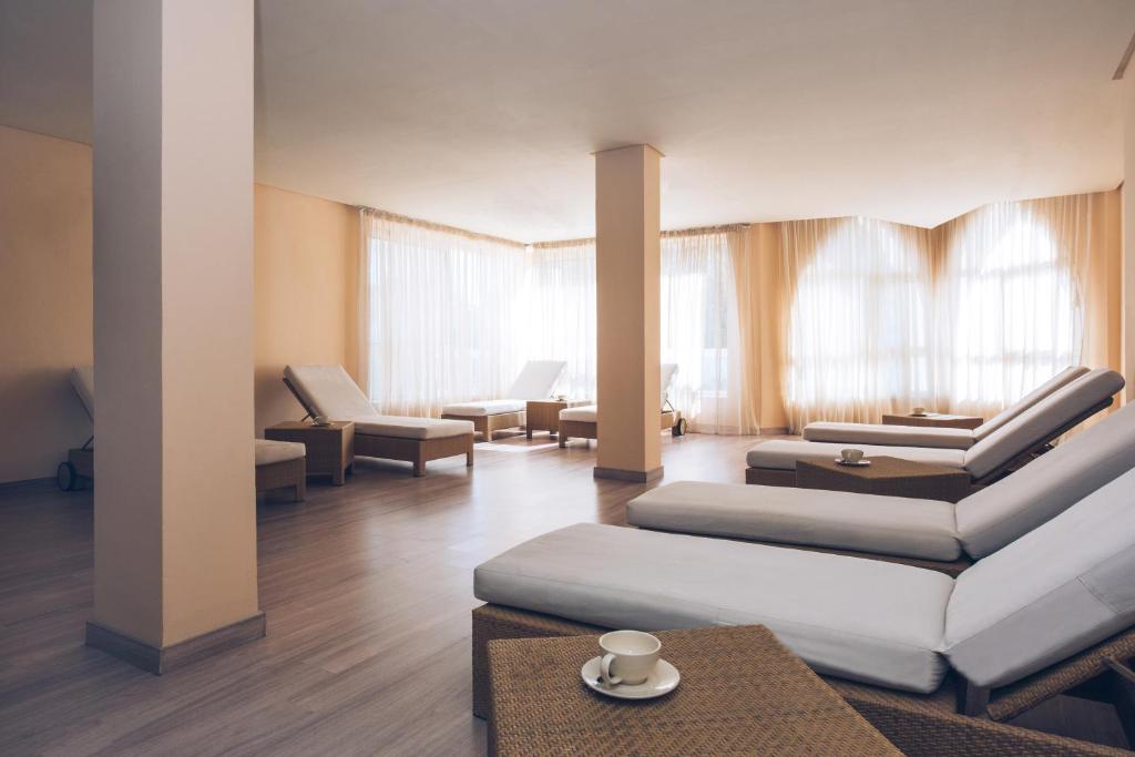 Relaksējoša spa telpa ar komfortabliem atpūtas krēsliem un dabiski apgaismotiem logiem, radot mierīgu atmosfēru. Uz galda novietota krūze ar tēju, piešķirot papildus komfortu.